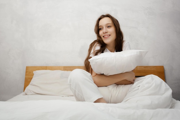 Young beautiful girl smiling while sitting in bed and hugging pillow Multiracial girl smiling to the camera while relaxing at the cozy bedroom Morning concept Stock photo