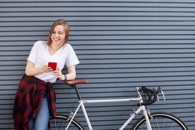 Young beautiful girl stands with a bicycle on the background of a wall and looks into the phone