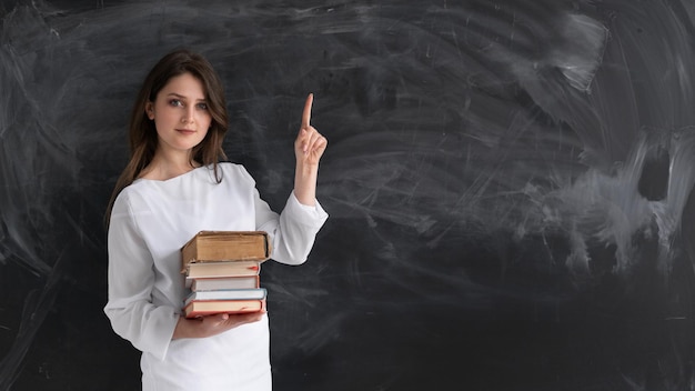 Young beautiful girl student stands against the background of a clean chalk board He holds a stack of textbooks in his hands The index finger points up