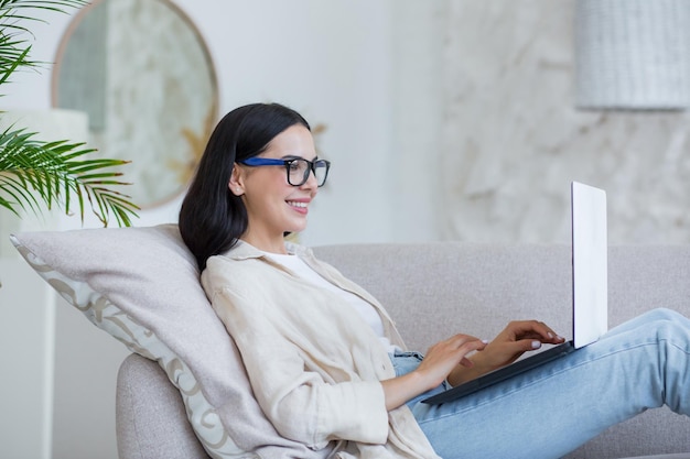 Young beautiful girl studying online at home sitting on sofa in living room brunette in glasses