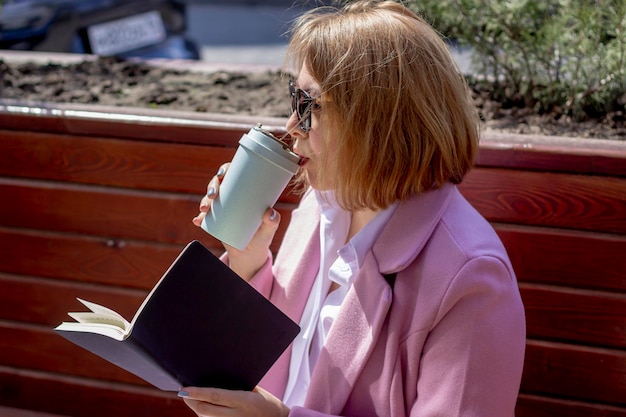 A young beautiful girl in sunglasses drinks coffee from an ecofriendly thermocup and holds a notebook while sitting on a bench outside in warm weather