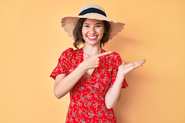 Young beautiful girl wearing summer hat amazed and smiling to the camera while presenting with hand and pointing with finger.