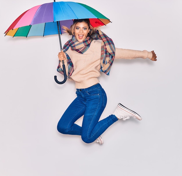 Young beautiful girl wearing winter clothes and scarf smiling happy Jumping with smile on face holding colorful umbrella over isolated white background