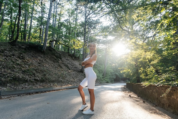 A young beautiful girl in white clothes poses before a running workout on a road in a dense forest during sunset Healthy lifestyle and running in the fresh air Youth