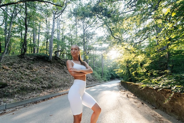 A young beautiful girl in white clothes poses before a running workout on a road in a dense forest during sunset Healthy lifestyle and running in the fresh air Youth