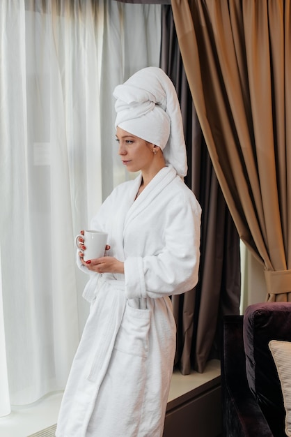 A young beautiful girl in a white coat is standing and drinking coffee near the window in her hotel room Rest and travel Hotel recreation and tourism