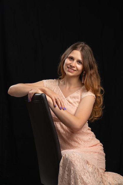 A young beautiful girl with dark long hair in a light pink dress sits on a black chair black backgroundStudio shooting