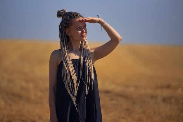 A young beautiful girl with dreadlocks stands in the middle of a field with dry grass and looks into...