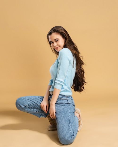 Young beautiful girl with long curly hair in a blue sweatshirt and blue jeans stands on one knee on a pastel orange.