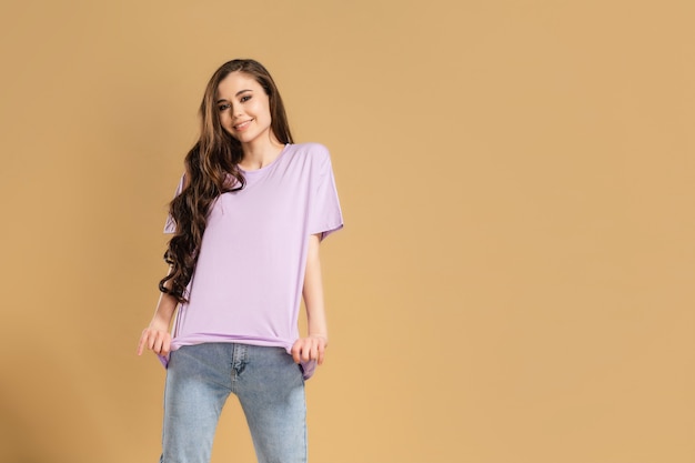 Young beautiful girl with long curly hair in a lilac T-shirt and blue jeans on a pastel orange.