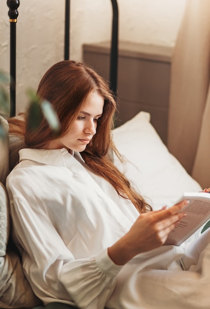 A young beautiful girl with long hair in a white shirt sits on the sofa and reads a book. Vertical photo