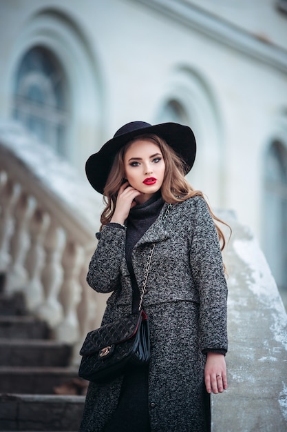 Young beautiful girl with perfect make-up, red lips, wearing a black hat and grey coat, black dress