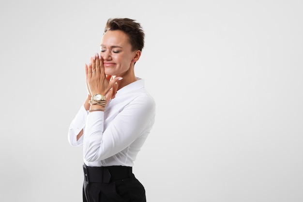 A young beautiful girl with short dark hair, makeup in a white shirt, black pants, with a wristwatch, bracelet and white manicure prays and smile