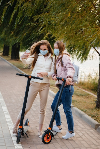 Young beautiful girls in masks ride in the Park on an electric scooter on a warm autumn day and take selfies. Walk in the Park.