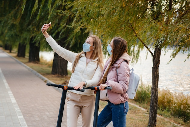 Young beautiful girls in masks ride in the Park on an electric scooter on a warm autumn day and take selfies. Walk in the Park.