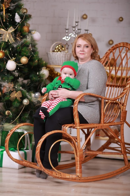 Young beautiful grandmother with her grandson in elf costume sitting in rocking chair near christmas tree.