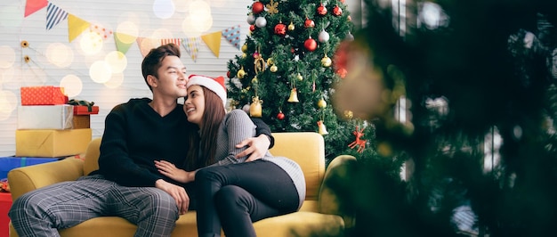 Young beautiful happy Asian woman wearing a Santa Claus hat sitting together with her boyfriend on the sofa at home with a Christmas tree in the background Image with copy space