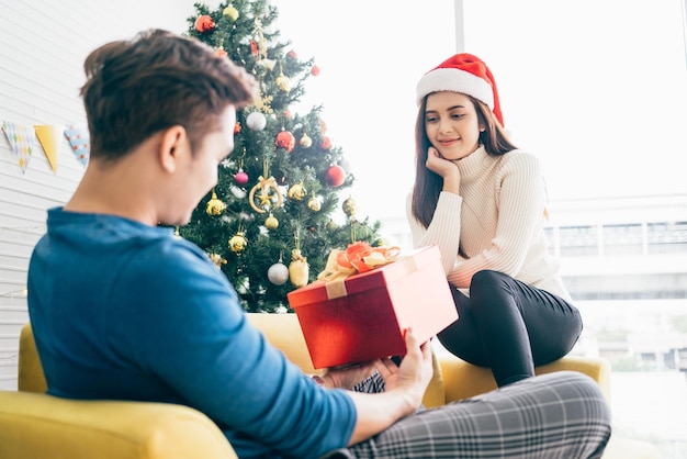 Young beautiful happy Asian woman wearing a Santa Claus hat surprises her boyfriend with gift at home with a Christmas tree in the background Celebrate the boxing day concept Photo with copy space