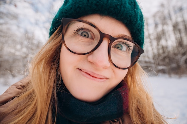 Young beautiful happy girl in green winter knitted hat takes selfie in winter forest. Looking at camera and smile. Travel and active life concept. Outdoors