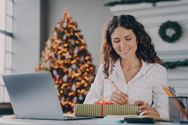 Young beautiful happy italian business woman signs prepared xmas gift box for client in office