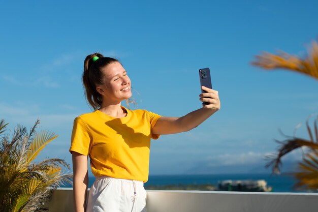 Young beautiful happy woman blogger is taking selfie picture of herself on frontal camera to social media of smartphone cell mobile phone smiling waving her hand having video call conversation