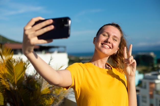 Young beautiful happy woman blogger is taking selfie picture of herself on frontal camera to social media of smartphone cell mobile phone smiling waving her hand having video call conversation