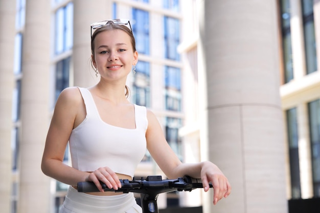 Young beautiful happy woman pretty attractive girl teenager is riding electro scooter in the city at summer sunny day in white clothes smiling in sunglasses Electric eco sharing transport