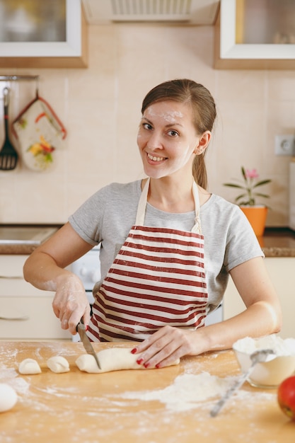 The young beautiful happy woman sitting at a table, cuts a dough with a knife into pieces and going to prepare a cakes in the kitchen. Cooking home. Prepare food.