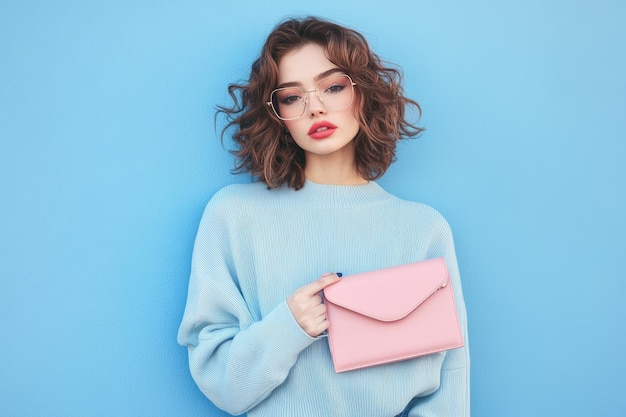 Young beautiful hipster woman posing against blue wall holding pink trendy clutch in hand