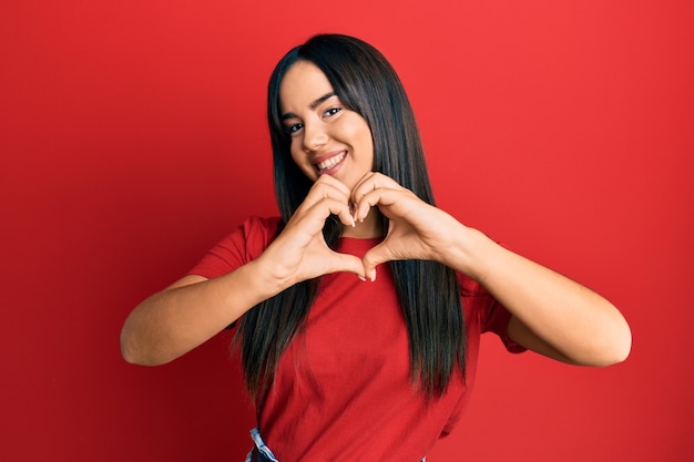 Young beautiful hispanic girl wearing casual red tshirt smiling in love doing heart symbol shape with hands. romantic concept.