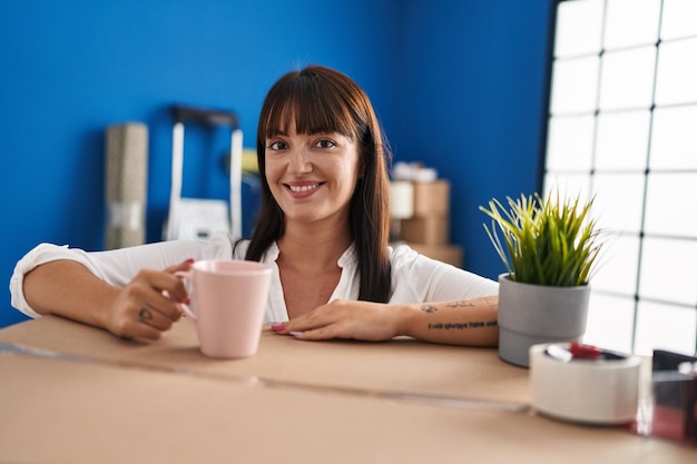 Young beautiful hispanic woman drinking coffee leaning on package at new home