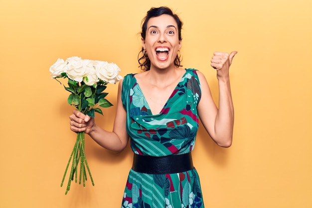 Young beautiful hispanic woman holding flowers pointing thumb up to the side smiling happy with open mouth