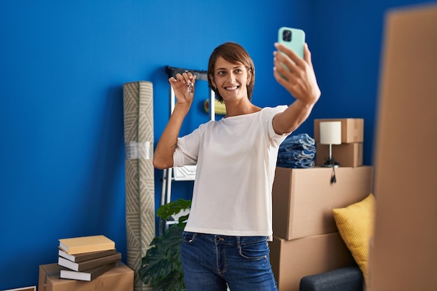 Young beautiful hispanic woman make selfie by smartphone holding key at new home