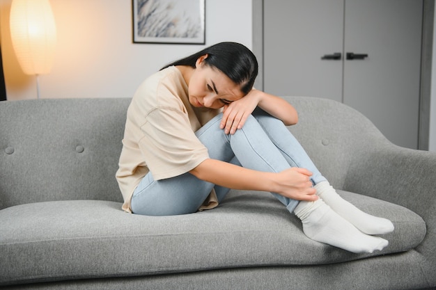 Young beautiful hispanic woman in painful expression holding her belly suffering menstrual period pain lying sad on home bed having tummy cramp in female health concept