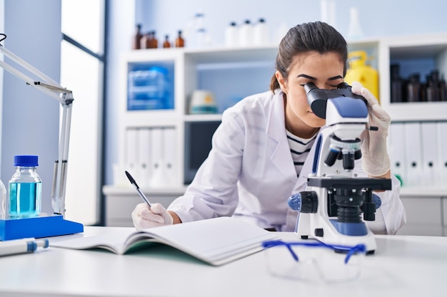 Young beautiful hispanic woman scientist using microscope writing on notebook at laboratory