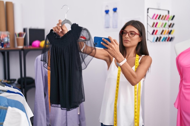 Young beautiful hispanic woman tailor make photo by smartphone to t shirt at tailor shop