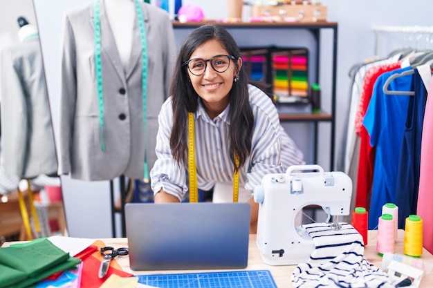 Young beautiful hispanic woman tailor smiling confident using laptop at clothing factory