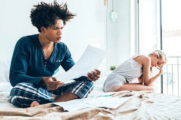 Young beautiful interracial couple in bed stressed with financial problems doing calculations with paper workxA