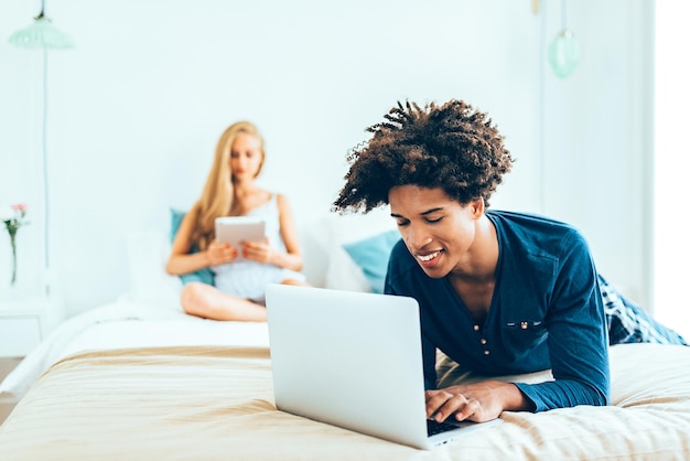 Young beautiful interracial couple relaxed lying down in bed on the computer and tablet