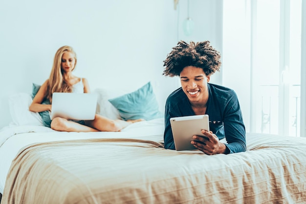 Young beautiful interracial couple relaxed lying down in bed on the computer and tablet