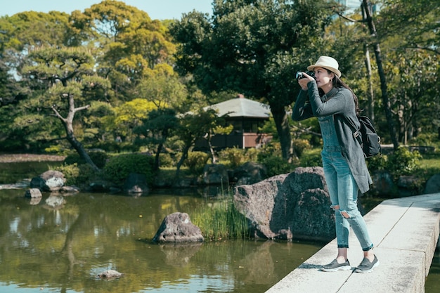 young beautiful lady photographer takes pictures of landscape in park. full length of elegant woman travel backpacker with camera stand on stone path above lake surrounding by green plant in spring