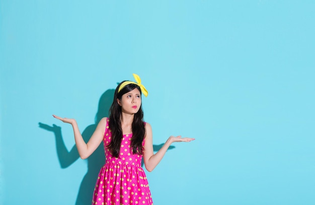 young beautiful lady showing confused emotion standing on blue background and wearing summer dress making choosing posing looking at empty area.