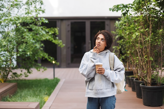 Young beautiful lady with dark curly hair standing with backpack and cup of coffee to go in hand while dreamily 