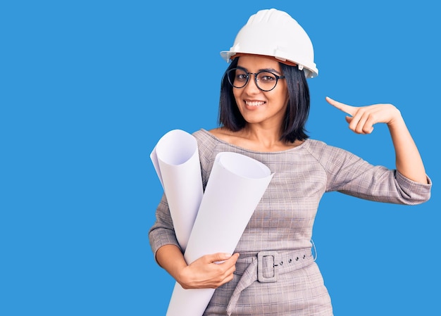 Young beautiful latin woman wearing architect hardhat and glasses holding blueprints pointing finger to one self smiling happy and proud