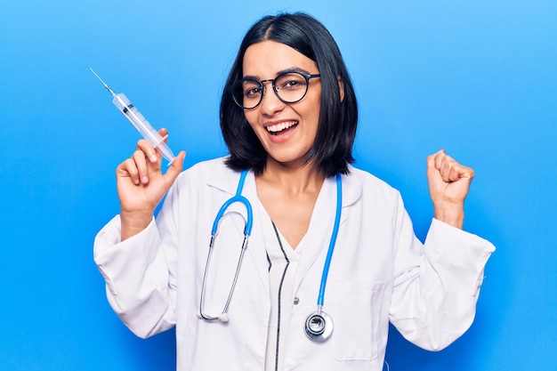 Young beautiful latin woman wearing doctor stethoscope holding syringe screaming proud celebrating victory and success very excited with raised arms