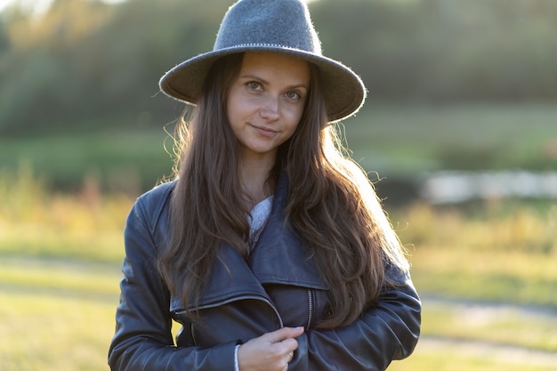 A young, beautiful long-haired woman in a hat smiles, looks at the camera in the park in the bright glow of sunset rays