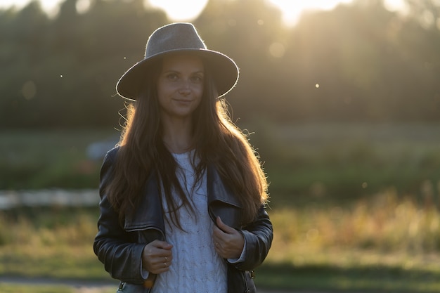 A young, beautiful long-haired woman in a hat smiles, looks at the camera in the park in the bright glow of sunset rays