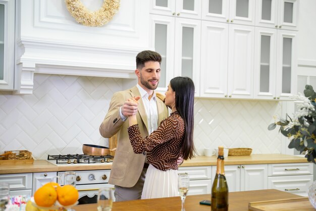 young beautiful married couple in festive wear dancing in light stylish kitchen near table