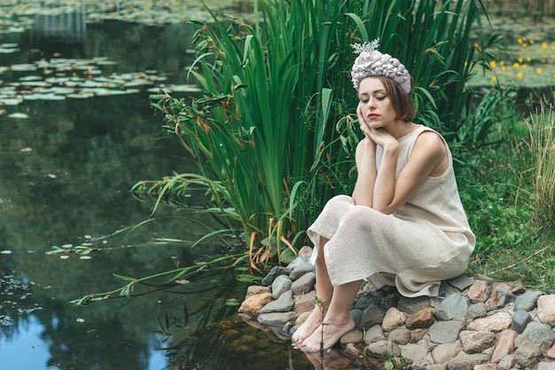 Young beautiful mermaid in seashells crown is sitting on the water backgroundMermaid cosplay