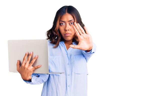 Young beautiful mixed race woman working using computer laptop with open hand doing stop sign with serious and confident expression defense gesture